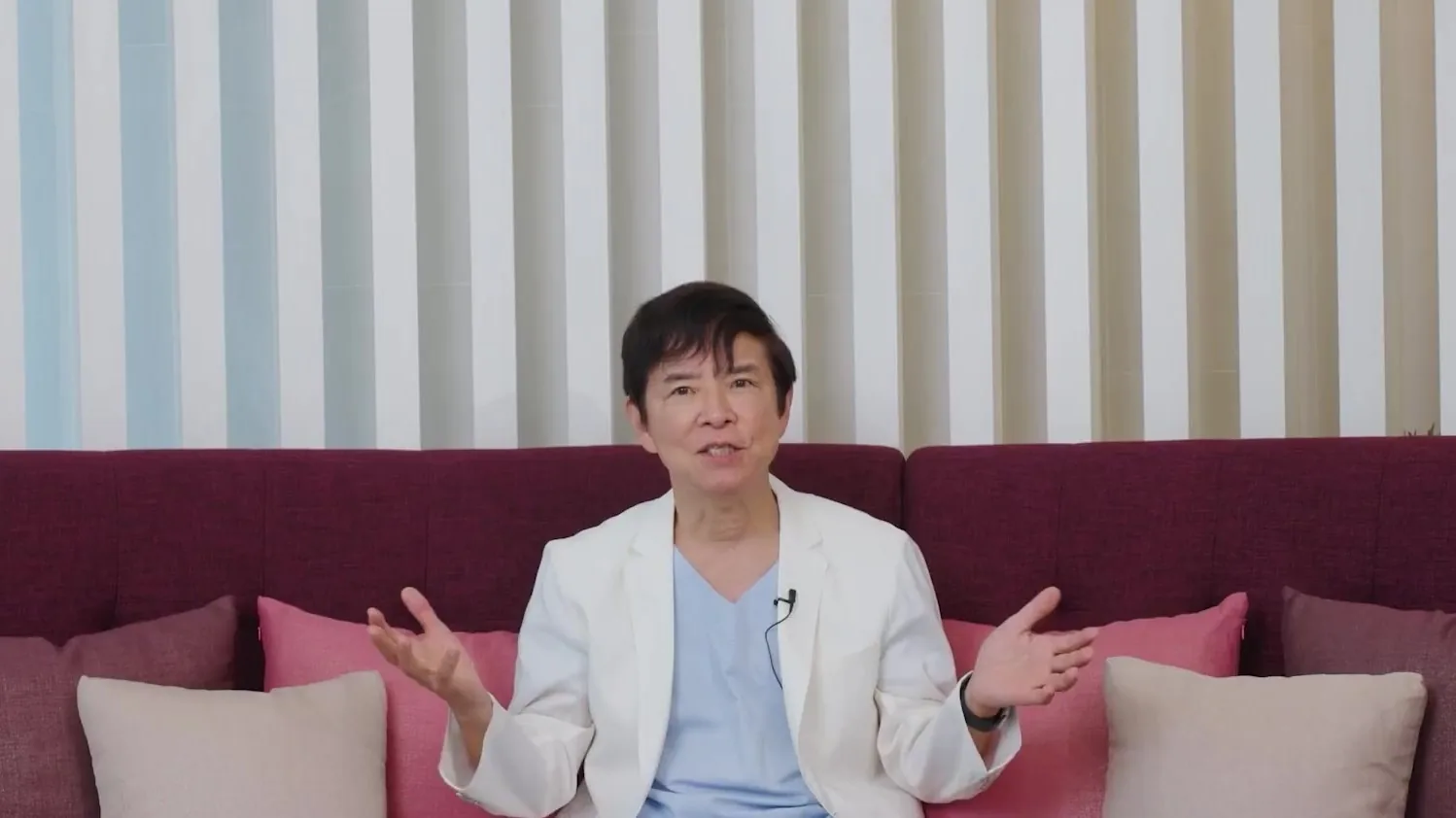 흰 재킷을 입은 의사가 소파에 앉아 두 손을 펴고 설명하는 모습